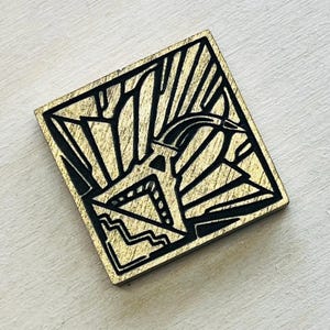 May include: Square wooden pin with a gold finish and black Art Deco design. The design features geometric shapes and lines, including a stylized bird or animal figure. The pin is likely for decorative purposes.