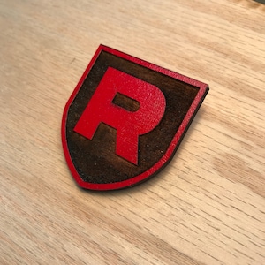 May include: A wooden shield-shaped cutout with a red letter "R" in the center. The shield is stained dark brown and has a red outline.