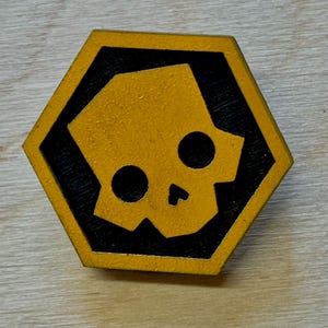 Pode incluir: Um broche hexagonal com uma silhueta de caveira amarela centrada num fundo preto. O design da caveira tem duas órbitas oculares circulares e uma pequena abertura nasal triangular. A peça é emoldurada por uma borda amarela espessa.
