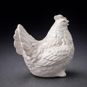 May include: A ceramic hen figurine in a light gray color. The hen is depicted in a side profile, with detailed feather textures and a small comb. The figurine is set against a dark background.