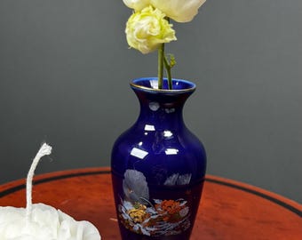 May include: A cobalt blue vase with gold trim holds a white and yellow flower. The vase has a decorative floral design. A white, rose-shaped candle sits on a red table.