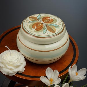 May include: A cream-colored ceramic jar with a lid featuring a floral design in brown, teal, and beige. The jar has horizontal brown and teal stripes. A white rose-shaped candle and white crocus flowers are also in the image.