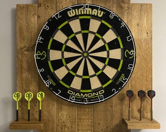 Op de afbeelding: Een houten dartbord wandbevestiging met een zwart-wit dartbord met de merknaam "Winmau" en de tekst "Diamond Plus" erop gedrukt. Het dartbord is gemonteerd op een rustieke houten wandbevestiging met twee planken voor darts. Er zijn groene en rode darts op de planken.