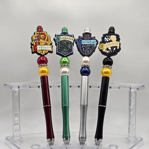 Op de afbeelding: Vier Harry Potter-huis-thema-pennen met hun respectievelijke wapenschilden bovenop. De pennen zijn rood, groen, zilver en zwart. De wapenschilden zijn Griffoendor, Zwadderich, Ravenklauw en Huffelpuf.