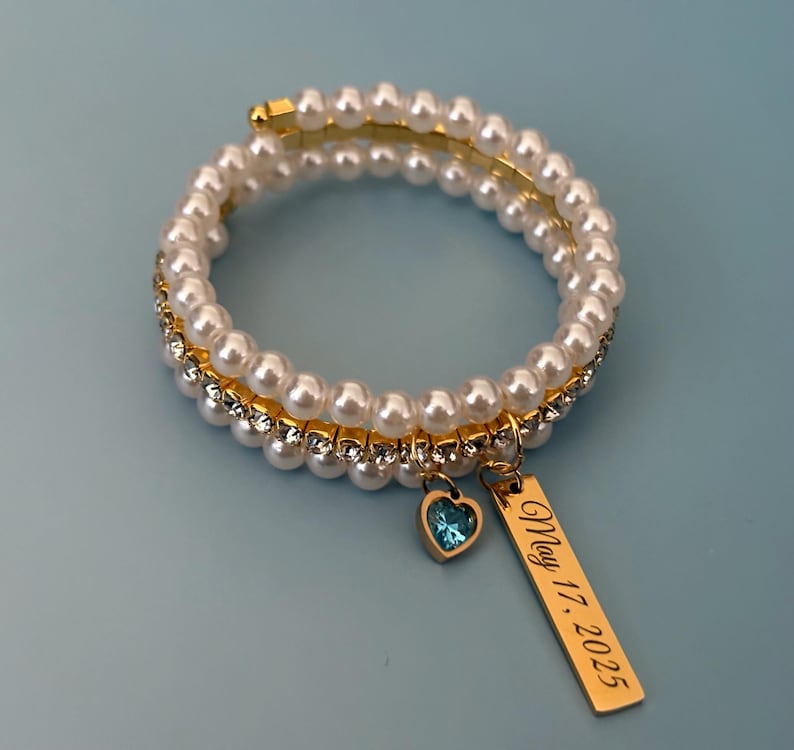 Pu&ograve; includere: Un braccialetto a spirale con perle finte, una fascia di strass trasparenti e una fascia in metallo dorato. Ha un piccolo ciondolo a forma di cuore blu e un ciondolo rettangolare dorato con l'iscrizione "May 17, 2025".