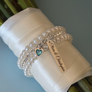 Pu&ograve; includere: Un bouquet avvolto in un nastro di raso bianco, adornato da braccialetti di perle e strass. Un'etichetta d'argento con i nomi "Olivia & Christopher" pende da un braccialetto, insieme a un ciondolo a forma di cuore con una gemma blu.