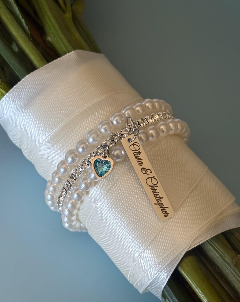 Pu&ograve; includere: Un bouquet avvolto in un nastro di raso bianco, adornato da braccialetti di perle e strass. Un'etichetta d'argento con i nomi "Olivia & Christopher" pende da un braccialetto, insieme a un ciondolo a forma di cuore con una gemma blu.