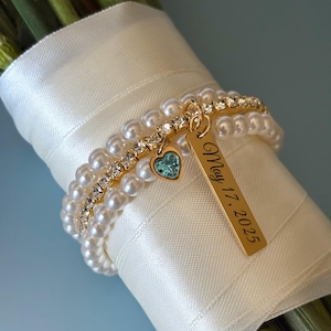 May include: A bouquet of flowers wrapped in white satin ribbon, adorned with a gold bracelet featuring pearls, rhinestones, and a teal heart charm. A gold tag reads "May 17, 2025".