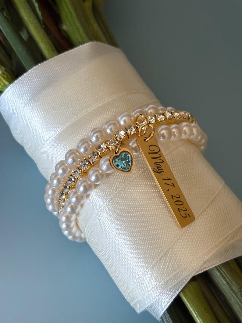 Pu&ograve; includere: Un bouquet di fiori avvolto in un nastro di raso bianco, adornato da un braccialetto d'oro con perle, strass e un ciondolo a forma di cuore turchese. Un'etichetta dorata recita "May 17, 2025".