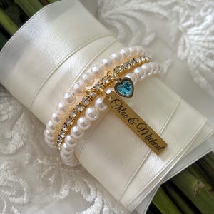 Pu&ograve; includere: Un bouquet da sposa avvolto in un nastro di raso avorio, adornato da braccialetti di perle e strass. Un'etichetta dorata con i nomi "Chloe & Michael" pende da un braccialetto con un ciondolo a forma di cuore blu. Lo sfondo &egrave; in tessuto di pizzo bianco.