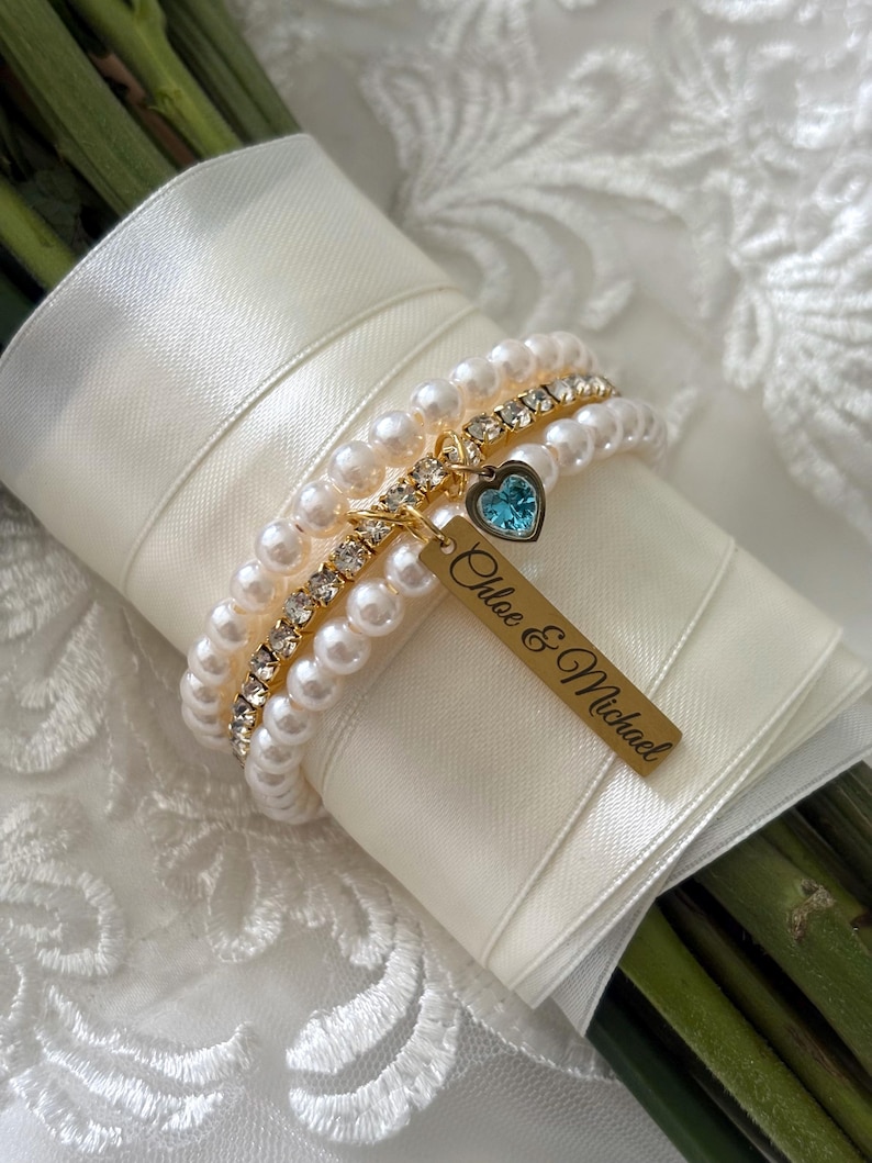Pu&ograve; includere: Un bouquet da sposa avvolto in un nastro di raso avorio, adornato da braccialetti di perle e strass. Un'etichetta dorata con i nomi "Chloe & Michael" pende da un braccialetto con un ciondolo a forma di cuore blu. Lo sfondo &egrave; in tessuto di pizzo bianco.
