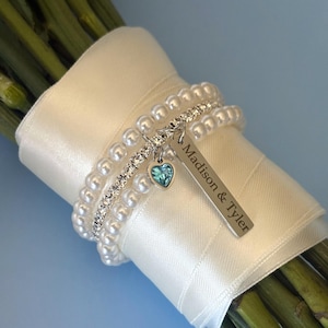 Pu&ograve; includere: Un bouquet di steli verdi avvolto con un nastro di raso bianco e adornato con un braccialetto di perle e strass. Un'etichetta argentata con la scritta "Madison & Tyler" pende da un piccolo ciondolo a forma di cuore con una gemma blu.