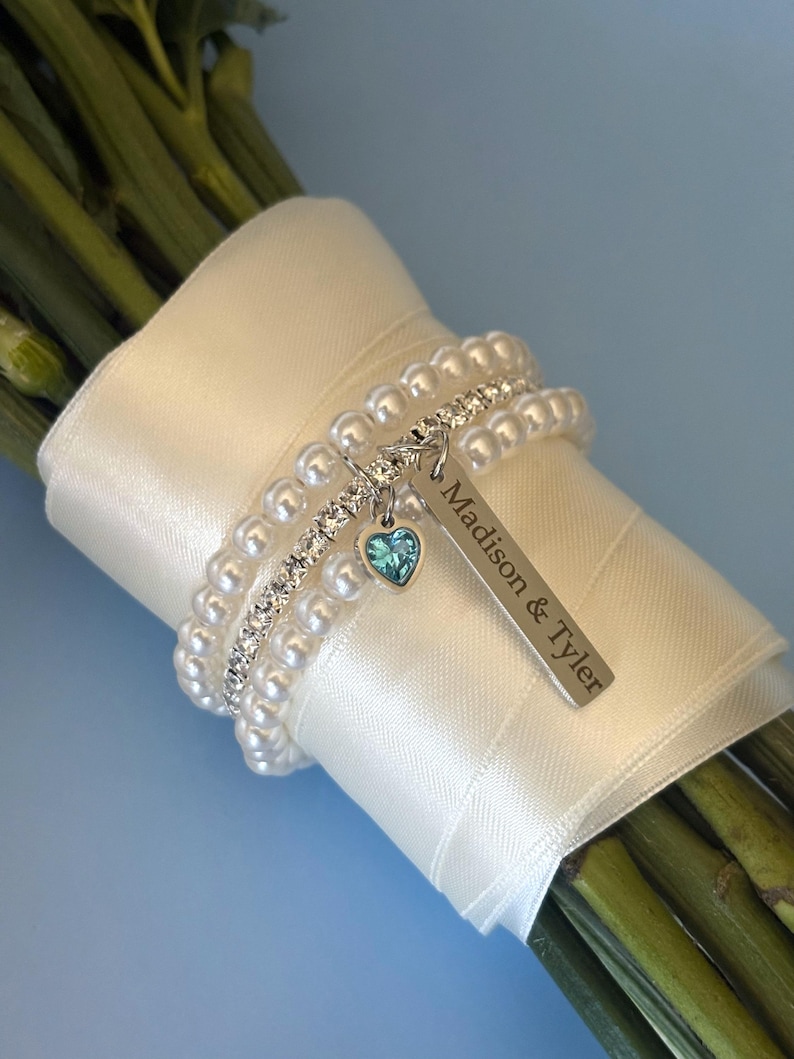 Pu&ograve; includere: Un bouquet di steli verdi avvolto con un nastro di raso bianco e adornato con un braccialetto di perle e strass. Un'etichetta argentata con la scritta "Madison & Tyler" pende da un piccolo ciondolo a forma di cuore con una gemma blu.