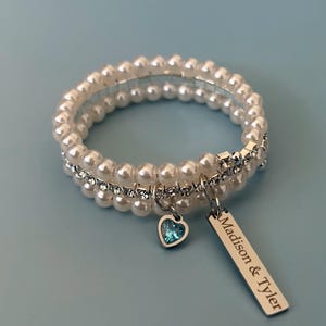 Pu&ograve; includere: Un braccialetto di perle bianche con una fascia argentata e un ciondolo a forma di cuore blu. Il braccialetto ha un'etichetta argentata con la scritta "Madison & Tyler". Il braccialetto &egrave; su uno sfondo azzurro.