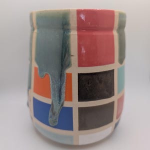 May include: A ceramic mug with a grid pattern of colorful squares in red, orange, blue, black, and teal. The mug has a cylindrical shape with a light-colored rim and base. Teal glaze drips down the side, adding a unique texture.