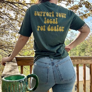 May include: Green t-shirt with the text "SUPPORT YOUR LOCAL POT DEALER" in yellow lettering. The person is wearing light blue jeans. A green ceramic mug sits on a wooden railing.