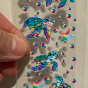 May include: A sheet of colourful sea turtle stickers with rainbow accents. The stickers feature turtles, starfish, and bubbles in shades of blue, green, pink, and yellow. The stickers are on a clear backing.