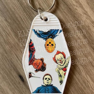 May include: White motel-style keychain featuring horror movie characters. The keychain includes images of a masked figure, a clown, and other iconic horror figures. The keychain has a silver metal ring.
