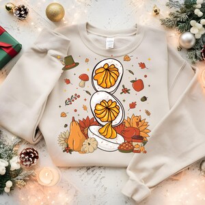 Könnte beinhalten: Ein cremefarbenes Sweatshirt mit einem Thanksgiving-Motiv. Das Design zeigt ein gefülltes Ei, Kürbisse, Herbstblätter und einen Truthahn. Das Sweatshirt ist von festlichen Dekorationen umgeben, darunter ein verpacktes Geschenk, Tannenzapfen und Ornamente.