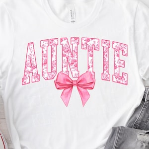 May include: White t-shirt with the word "AUNTIE" in pink floral block letters and a pink bow graphic. The shirt is paired with gray denim jeans and white sneakers.