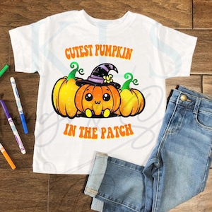 Op de afbeelding: Wit T-shirt met de tekst "Cutest Pumpkin in the Patch" in oranje. Het shirt heeft een afbeelding van drie pompoenen, waarvan één een heksenhoed draagt. Een blauwe jeans is naast het shirt opgevouwen.