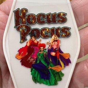 May include: White keychain with the words "Hocus Pocus" in brown and gold lettering. The keychain features a colorful graphic of three witches in red, green, and purple dresses. A silver key ring is attached to the top.