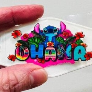 Może przedstawiać: Biały, owalny brelok do kluczy z napisem "OHANA" w kolorowych, stylizowanych literach. Niebieska postać z kreskówki znajduje się nad literami, otoczona tropikalnymi kwiatami i liśćmi. Brelok jest trzymany w dłoni.