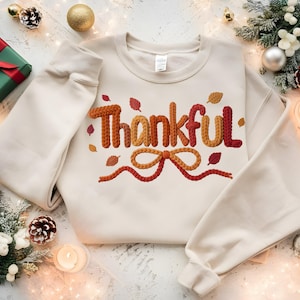 Könnte beinhalten: Cremefarbenes Sweatshirt mit dem Wort "Thankful" in einem strukturierten Design im Herbststil. Das Design umfasst orange, rote und braune Blätter und eine Schleife. Das Sweatshirt ist von Weihnachtsdekorationen umgeben, darunter ein Geschenk und Ornamente.