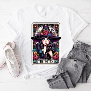 May include: White t-shirt with a colorful illustration of a witch holding a black cat. The illustration is framed in a tarot card style with the text "The Witch" at the bottom.