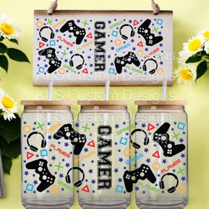 May include: Three clear glass tumblers with lids and straws. Each tumbler has a black and white gamer design with the word "GAMER" printed vertically on the side. The design includes game controllers, headphones, stars, and other gaming symbols in various colors.