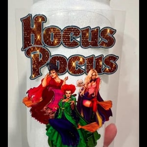 May include: A clear glass jar with a wooden lid, featuring the text "Hocus Pocus" in a glittery font. The jar is decorated with an image of three witches in colorful attire, holding brooms. The jar is held by a hand.