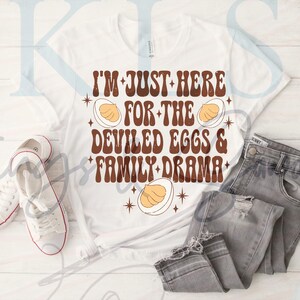 Könnte beinhalten: Weißes T-Shirt mit dem Text "I'M JUST HERE FOR THE DEVILED EGGS & FAMILY DRAMA" in braunen Buchstaben. Das Design beinhaltet Illustrationen von gefüllten Eiern. Das Shirt wird mit grauen Jeans und weißen Turnschuhen kombiniert.