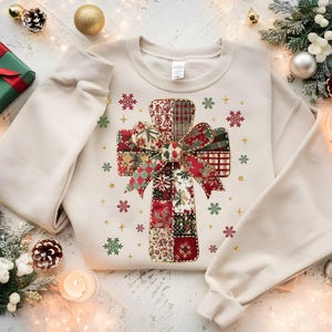 May include: A cream-colored sweatshirt featuring a cross design made of patchwork fabric in red, green, and white patterns. The cross is adorned with a bow and surrounded by snowflakes and gold stars. The sweatshirt is surrounded by Christmas decorations.