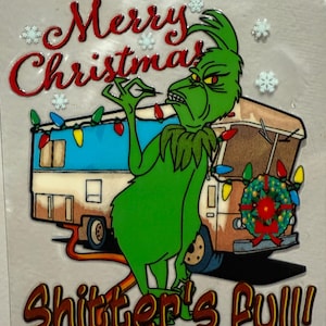 May include: A green Grinch character with an angry expression stands in front of a camper decorated with Christmas lights and a wreath. The text "Merry Christmas" and "Shitter's Full!" are displayed in red and brown.