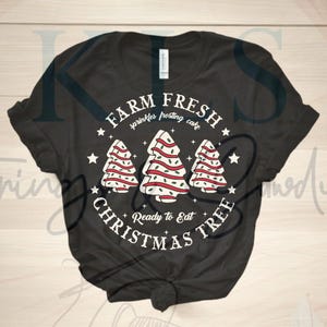 May include: Dark gray t-shirt with a festive design. The graphic features three Christmas tree illustrations with red ribbon and sprinkles, surrounded by the text "FARM FRESH sprinkles frosting cake CHRISTMAS TREE Ready to Eat."