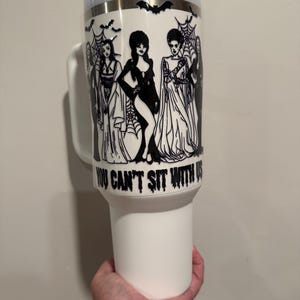 May include: White tumbler with a handle, a white lid, and a straw. The tumbler features a black and white graphic of several women in gothic attire, with the text "YOU CAN'T SIT WITH US". A bat design is also present.