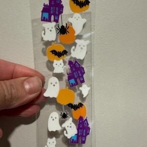 Puede incluir: Una lámina transparente de pegatinas con temática de Halloween con casas encantadas moradas, calabazas naranjas con murciélagos negros, fantasmas blancos y arañas negras. Las pegatinas están sujetas por una mano.