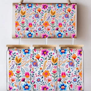 May include: Three glass jars with wooden lids and a matching wooden sign, all adorned with a vibrant floral pattern in orange, pink, blue, and yellow. The jars have white straws.