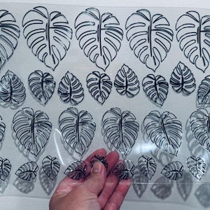 May include: Clear sheet of tropical leaf stickers with black outlines. The sheet contains various sizes of monstera leaves. A hand holds a few of the stickers, showing their transparent nature. The stickers are designed for crafting and decoration.