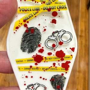 以下が含まれることがあります： 犯罪現場のグラフィックが描かれた白いキーホルダー。指紋、手錠、赤い飛沫、そして「POLICE LINE - DO NOT CROSS」と書かれた黄色のテープがデザインに含まれています。