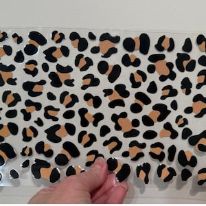 Pode incluir: Uma folha de autocolantes com estampado de leopardo. Os autocolantes têm um fundo transparente e apresentam manchas pretas e bege de várias formas e tamanhos. Uma mão segura um dos autocolantes.