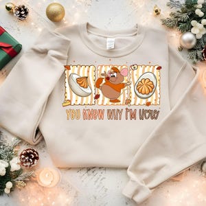 May include: Cream-colored sweatshirt with a graphic featuring a cartoon mouse, a deviled egg, and the text "YOU KNOW WHY I'M HERE." The design has a retro style with orange and brown accents. Festive decorations surround the sweatshirt.