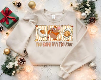 May include: Cream-colored sweatshirt with a graphic featuring a cartoon mouse, a deviled egg, and the text "YOU KNOW WHY I'M HERE." The design has a retro style with orange and brown accents. Festive decorations surround the sweatshirt.
