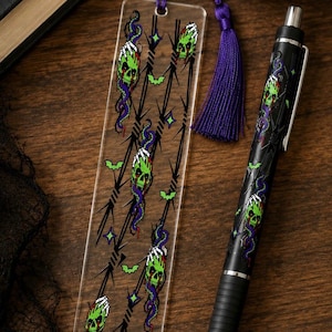 Puede incluir: Un marcapáginas rectangular transparente con un patrón de calaveras verdes, serpientes moradas, alambre de espino negro, pequeños murciélagos verdes y estrellas, con una borla morada, junto a un bolígrafo retráctil negro a juego.