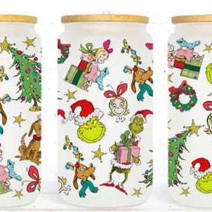 May include: Three white glass can-shaped tumblers with gold-colored lids, decorated with colorful cartoon characters and Christmas-themed elements. The design includes the Grinch, Cindy Lou Who, Max the dog, Christmas trees, and wrapped gifts.