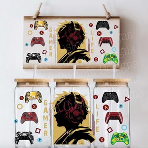 May include: Three glass tumblers with wooden lids and straws, featuring a gamer theme. Each tumbler has a design of a gamer silhouette with headphones and game controllers. A wooden sign with the same design is above the tumblers.