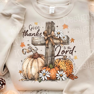 May include: Cream-colored sweatshirt featuring a graphic design with a wooden cross, pumpkins, and daisies. The text "Give thanks to the Lord" is displayed. The design incorporates fall colors and elements, perfect for autumn.