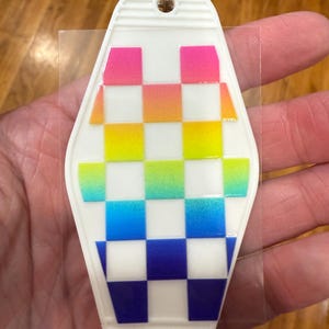 Könnte beinhalten: Weißer Schlüsselanhänger im Motel-Stil mit einem Regenbogen-Karomuster. Das Karomuster wechselt von Pink zu Orange, Gelb, Grün und Blau. Der Schlüsselanhänger hat oben einen Metallring zur Befestigung.