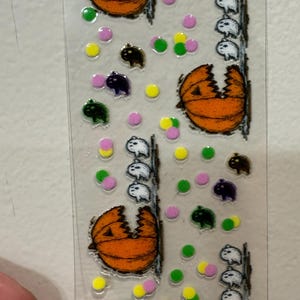Puede incluir: Hoja de pegatinas transparente con diseños de Halloween. Presenta calabazas naranjas, fantasmas blancos y pequeñas criaturas negras, verdes y moradas. Puntos de colores en amarillo, rosa y verde completan el aspecto festivo.