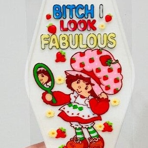 Puede incluir: Llavero de plástico blanco con una imagen de dibujos animados de una niña con sombrero de fresa y el texto "BITCH I LOOK FABULOUS". El llavero tiene un lazo de metal.